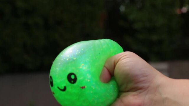 A hand squeezes a cute sparkly green dumpling stress toy with small ears and a kawaii face, set against a dark blurred background