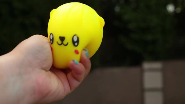 A hand squeezes a cute yellow round dumpling stress toy with small ears and a kawaii face, set against a dark blurred background