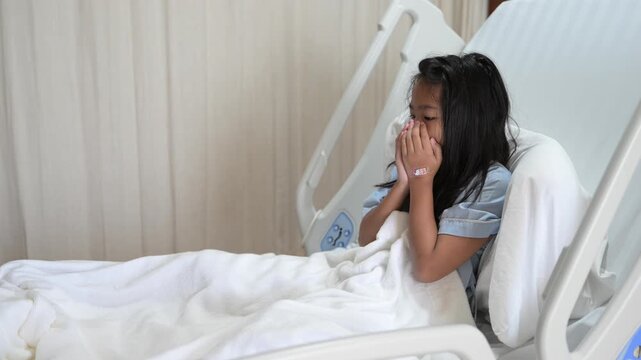 Sick young Asian child patient sitting in a modern hospital bed coughing heavily into her hands suffering from a severe respiratory illness during pediatric healthcare recovery inside a clinic.