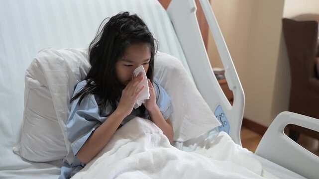 Sick young Asian child patient sitting in a modern hospital bed coughing heavily into her hands suffering from a severe respiratory illness during pediatric healthcare recovery inside a clinic.