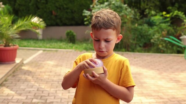 A boy holds a sparkly purple dumpling toy in his hands, squeezing its glittery squishy surface while standing outdoors in natural daylight