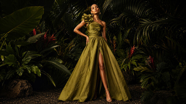 Elegant woman in a flowing olive gown posing in a dark tropical garden.