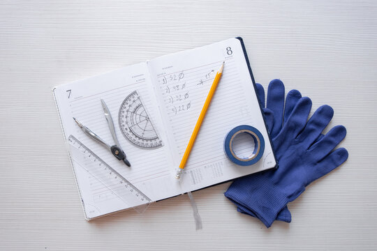 Engineering notes in a notebook, written in pencil. On top of the notebook are a measuring tape, dividers, and protractor, and underneath the notebook are a pair of construction gloves.