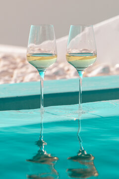 Two glasses of white wine stand beside a turquoise pool, their reflections shimmering in sunlight, evoking summer relaxation and Mediterranean elegance.