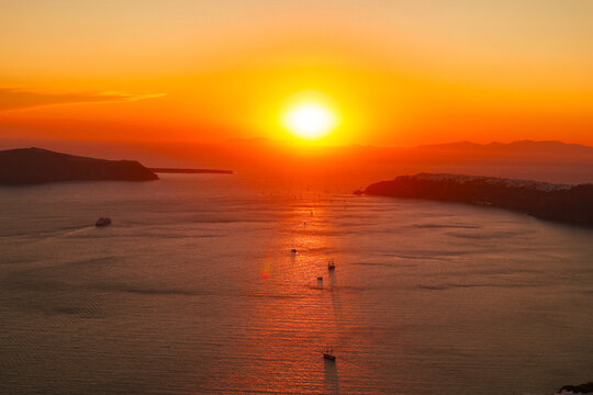 Golden red sunset over the Santorini caldera with boats gliding across shimmering water, capturing the island&rsquo;s tranquil Mediterranean beauty and vivid evening colors.