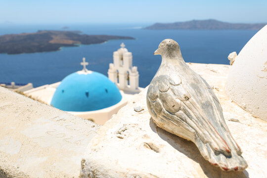 A weathered bird sculpture rests on a white wall overlooking the iconic blue‑domed church and caldera of Santorini, Greece, under bright Mediterranean sunlight.