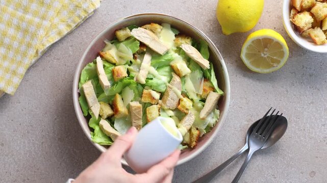 Overhead video of a hand pouring creamy Caesar dressing over a homemade Caesar salad with grilled chicken, Parmesan cheese and croutons. Flat lay cooking scene. Horizontal video.