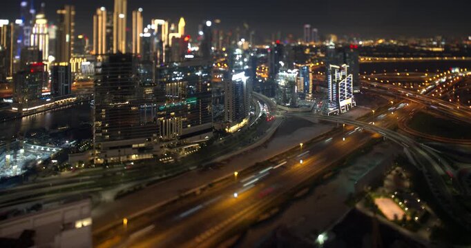 Dubai at night, car light trails run along the wide highway, lit towers and the water canal fill the frame, tilt shift lens turns the city into a miniature model, timelapse