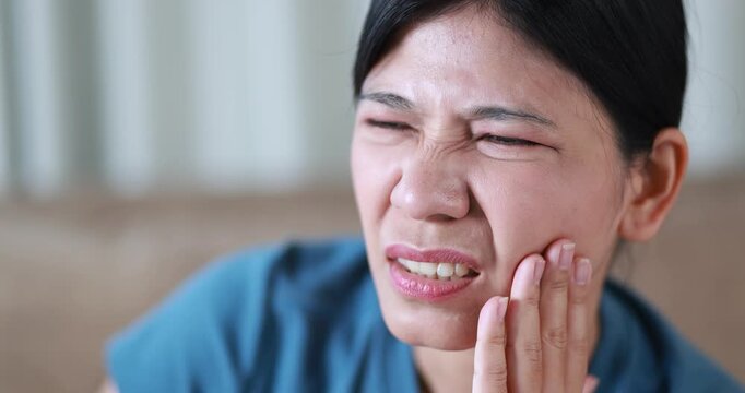 Woman suffering from toothache, holding her cheek in pain, tooth pain, dental problems, oral health, tooth sensitivity, cavity, dental treatment, and healthcare, close-up, Asian Thai people