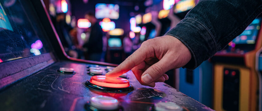 Hand pressing glowing red button on retro arcade cabinet in neon-lit game room