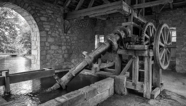 A black and white view of an old water-powered mill mechanism with large wooden gears and iron shafts inside a stone building by a river