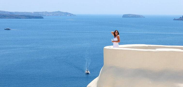 Woman standing on a white terrace overlooking the caldera and Aegean Sea in Oia, Santorini, Greece, with boats and volcanic islands in the distance.