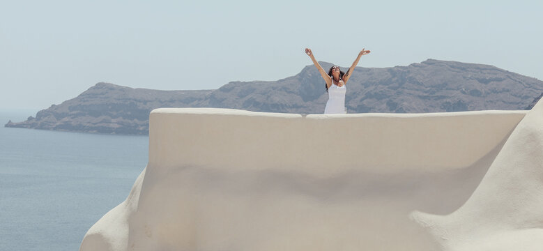 Woman in a white dress raising her arms in celebration and gratitude on a white terrace overlooking the caldera in Oia, Santorini, Greece.