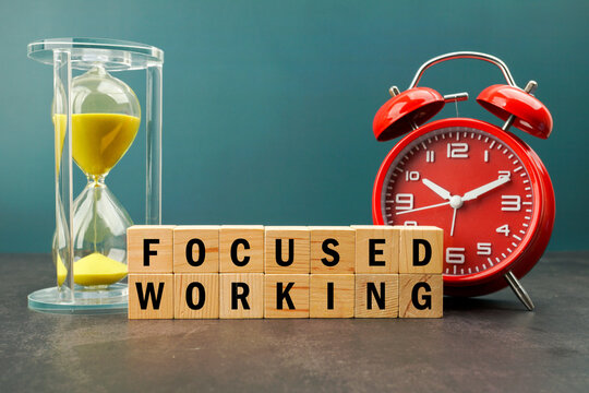 Hourglass and alarm clock behind wooden blocks spelling FOCUSED WORKING. Concept of deep work, productivity, concentration, time management and workplace efficiency.