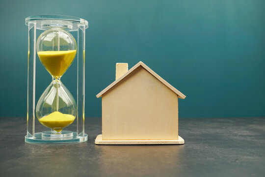 Wooden house model beside an hourglass on a dark surface. Concept of home ownership, mortgage planning, property investment, real estate timing and long term financial goals.