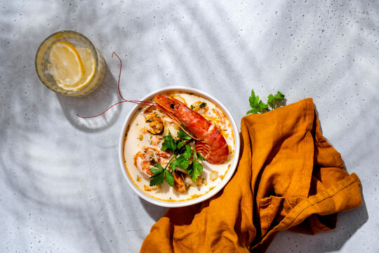 Seafood chowder soup with langoustine, shrimp, mussels with napkin, lemon drink on concrete surface. Overhead restaurant banner background, gourmet seafood culinary presentation concept.