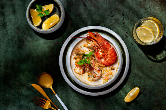 Creamy seafood chowder soup with jumbo prawn, mussels, and golden cutlery in hard light. Elegant restaurant layout, on dark green background with citrus drink.