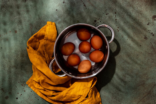 Brown raw chicken eggs submerged in water inside stainless steel pot, with ochre linen napkin on green background in hard sunlight.