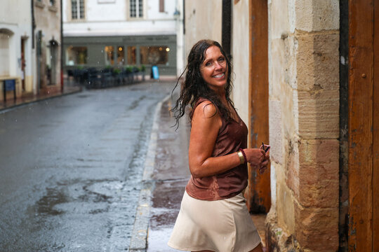 Woman standing in a rain shower on a historic street in Beaune, Burgundy, France, with wet pavement, stone architecture, and summer rainfall creating an authentic travel scene.