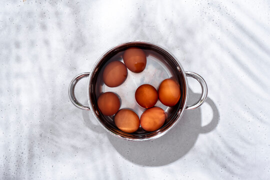 Top view of stainless steel pot filled with water and brown eggs, centered on light grey background under bright sunlight.