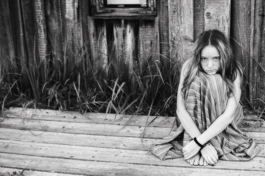 Black and white environmental portrait of a barefoot teenage girl wrapped in a striped blanket sitting alone on a weathered wooden porch outside a rustic house