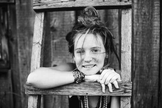 Black and white portrait of a smiling teenage girl leaning through a wooden ladder outdoors creating a playful and authentic childhood moment