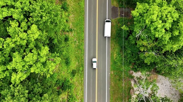 Aerial top-down view of an SUV overtaking a truck on a two-lane forest road. Drone footage of highway traffic, transportation, safe passing, and scenic woodland travel.