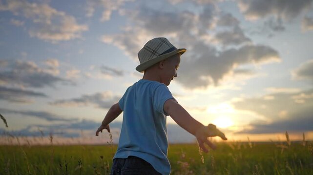 Happy boy stands in green field with arms outstretched at sunset. Little kid pretends to be flying plane during summer evening. Child wears hat while playing outdoors. Boy enjoys sunset in field.