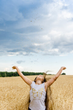 Happy little girl throwing wheat spikelets in air in golden field under blue sky