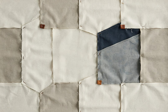 Fabric patchwork quilt made of square textile pieces in beige, cream, and dark blue, joined with visible pins on a light background
