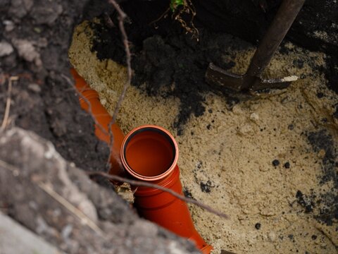 An orange plastic sewer pipe branch installation inside a deep dirt trench with sand bedding and a shovel.