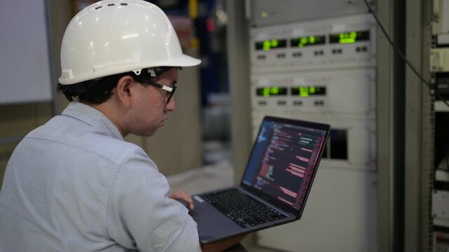 Focused professional male industrial engineer programs an advanced smart factory electrical control cabinet using automation software on a digital laptop inside a modern manufacturing plant.