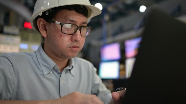 Focused professional Asian industrial engineer in a hard hat programs automation software on a laptop inside a modern smart factory control room managing production system network data.