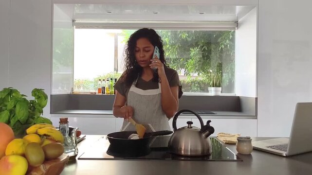 Hispanic woman talks on her smartphone while stirring food cooking in a cast iron skillet on a modern induction cooktop in a bright contemporary kitchen