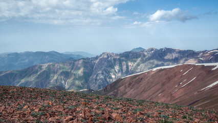 Rocky alpine foreground with sparse vegetation overlooking colorful mountain ridges and lingering snowfields under evening light. © Kolchanov