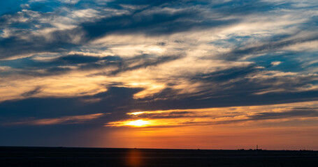 Fototapeta premium A dramatic sunset over a flat horizon. Vibrant orange and golden sunbeams break through layered clouds in a darkening blue sky. A scenic natural background for design and concepts.