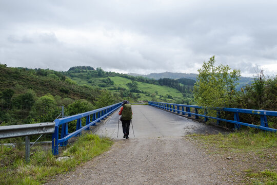 Pilgrim with backpack walking across a blue bridge on the Camino Primitivo trail in Asturias, Spain