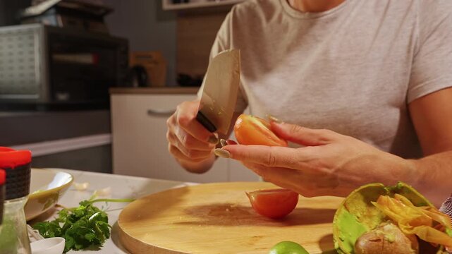 Hands Slice Fresh Tomato on a Wooden Board in Kitchen, With Avocado Limes and Cilantro Supporting Fresh Food Preparation