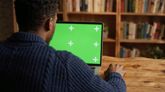 Man working on laptop with green screen chroma key display in cozy home library office for remote work and digital marketing projects