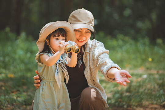 Happy Asian mother and her little daughter with binoculars exploring nature together in green park, cute toddler child learning outdoors on family summer vacation for early childhood education, parent