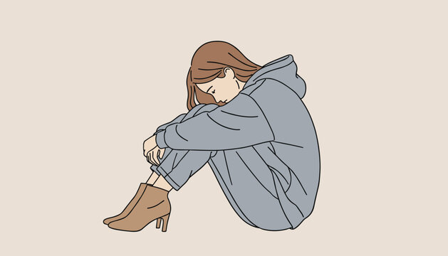 Lonely depressed woman wearing a grey hoodie sitting on the floor hugging her knees and hiding her face due to emotional distress or grief.