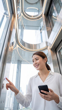 A smiling young Asian woman in a professional white blouse stands inside a futuristic modern glass elevator in a skyscraper, holding a smartphone while pressing a button to select her floor.