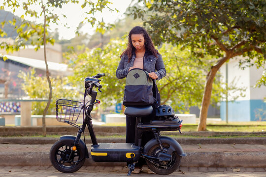 Young Black woman preparing her backpack before commuting on her electric bicycle, parked on a park sidewalk surrounded by trees.