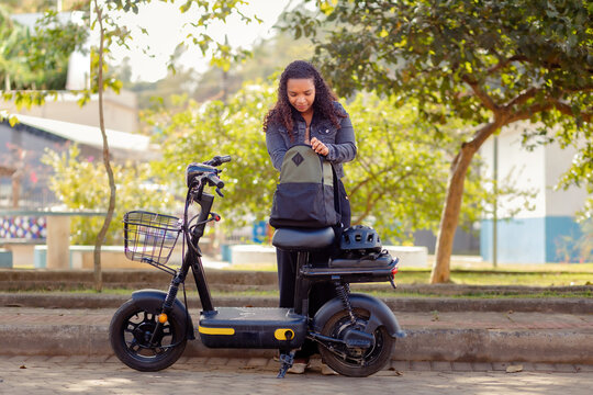 Young Black woman preparing her backpack before commuting on her electric bicycle, parked on a park sidewalk surrounded by trees.