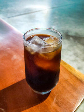 Iced Americano served in a tall glass at the edge of a wooden caf&eacute; table, featuring rich dark coffee, melting ice cubes, and a relaxed atmosphere for casual coffee moments.