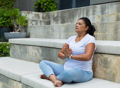 Senior woman sitting meditating on patio padded steps wearing white shirt blue leggings, copy space