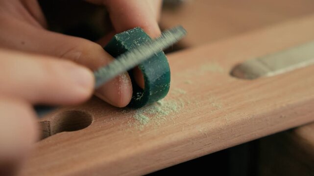 Close-up footage of a jeweler's hands holding a green wax ring model and filing it with a small needle file on a wooden bench pin. Fine wax shavings fly off as the artisan shapes the ring.