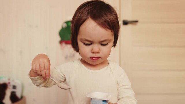 Little girl eating yogurt with a spoon at home and happily showing she likes the taste