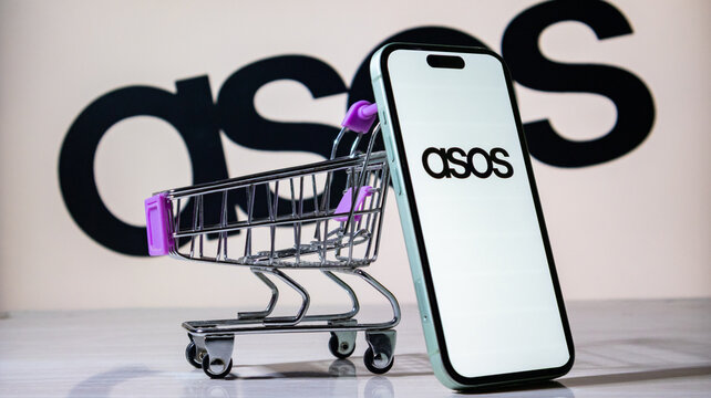 ASOS logo displayed on a smartphone with a miniature shopping cart in front of a blurry background.