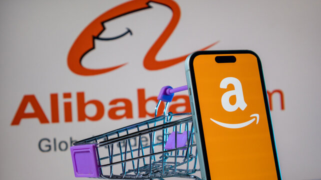 Miniature shopping cart next to smartphone with Amazon app logo on Alibaba logo background.